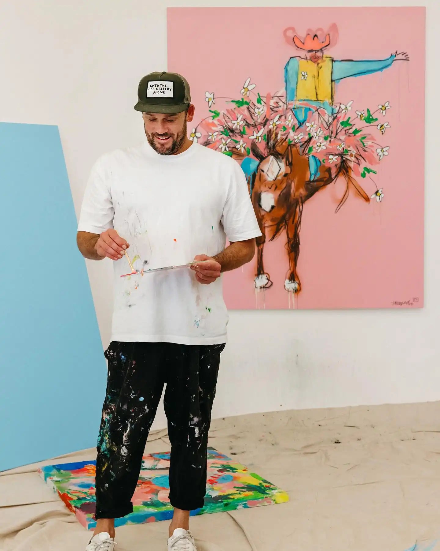 Jakey Pedro holding a piece of paper in front of a colorful painting of a horse and cowboy a pink background.